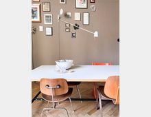 Esszimmer mit einem weißen Tisch, umgeben von Holzstühlen und einem Einzelstuhl in Orange; an einer braunen Wand hängen Bilderrahmen und eine schwenkbare weiße Lampe. Der Boden besteht aus hellem Parkett.