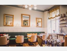 Ein gemütlicher Innenraum eines Cafés mit weiß getäfelten Wänden, grünen Polsterbänken und beigefarbenen Stühlen. An den Wänden hängen goldgerahmte Spiegel und Gemälde, während auf der rechten Seite ein Regal mit Teekannen und ein Samowar stehen; große Fenster lassen Tageslicht herein.