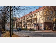 Straßenszene mit einem mehrstöckigen Wohnhaus im Stil des frühen 20. Jahrhunderts, das gelbe Putzfassaden und rote Backsteinbasis hat. Entlang der Straße stehen kahle Bäume und es sind parkende Autos sowie ein Fußgänger erkennbar.