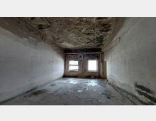 Ein leerer, heruntergekommener Raum mit abblätterndem Putz an den Wänden sowie Decken- und Wandbeschädigungen. Zwei Fenster an der Stirnwand lassen Tageslicht in den Raum, während der Boden Spuren von Feuchtigkeit und Abnutzung zeigt.