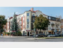 Eckgebäude mit mehrstöckigen Fassaden im Stil klassischer Wohnhäuser, kombiniert aus gelben und roten Backsteinflächen, mit Balkonen und Stuckverzierungen. Vor dem Gebäude stehen Bäume, parkende Autos und eine Ampel an einer Straßenkreuzung, daneben ein Schild mit der Aufschrift „Biergarten“.