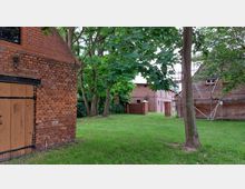 Ziegelgebäude mit rotem Backstein und Holztüren, umgeben von Bäumen und Grasflächen; im Hintergrund weitere Ziegelbauten und ein Baugerüst sichtbar.