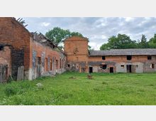 Backsteingebäude in einem verfallenen Zustand, mit teilweise eingestürztem Dach und beschädigten Fenstern, umgeben von einer grünen Wiese. Im Hintergrund sind Bäume und ein bewölkter Himmel zu sehen.
