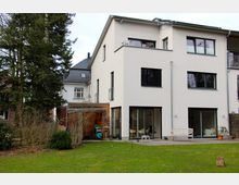 Modernes Wohnhaus mit drei Etagen und heller Putzfassade, großen Fenstern und teilweise bodentiefen Glasfronten. Im Vordergrund befindet sich ein gepflegter Rasen mit einigen Bäumen und Sträuchern, sowie eine Terrasse mit Holzmöbeln.