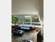 Wohnzimmer mit großen, bodentiefen Fenstern, die Blick auf eine grüne, bewaldete Umgebung bieten. Im Raum stehen ein schwarzes Ledersofa, ein niedriger Holztisch, ein Teppich und eine große Topfpflanze, ergänzt durch eine Stehlampe und helle Holzböden.