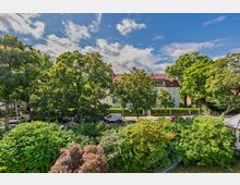 Blick über dichte, grüne Baumkronen auf eine ruhige Wohnstraße mit geparkten Autos und eine mehrstöckige Villa mit hellem Putz und rotem Ziegeldach. Im Hintergrund sind weitere Bäume und ein blauer Himmel mit Wolken zu sehen.