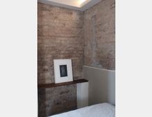 Ecke eines Raumes mit unverputzter Backsteinwand, einer weißen halbhohen Wandverkleidung und einer schmalen Holzablage, auf der ein gerahmtes Schwarz-Weiß-Bild steht. Im Vordergrund ist ein Teil eines Bettes mit weißer Decke erkennbar.