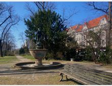 Ein Park mit einer steinernen Brunneninstallation im Vordergrund, die mit einer Statue verziert ist und Graffiti aufweist. Im Hintergrund sind Bäume und ein historisches Gebäude mit roten Dächern zu sehen.