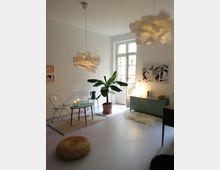 Helles Wohnzimmer mit minimalistischer Einrichtung, einschließlich eines runden Beistelltischs mit Kerzen, zwei transparenten Stühlen, einem Bananenbaum im Topf und einer grünen Kommode. Der Raum hat weiße Wände, große Fenster mit Doppeltüren und dekorative Hängeleuchten im organischem Design.