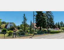 Wohnstraße mit Einfamilienhäusern, Vorgärten und Metallzäunen; im Vordergrund eine unbefestigte Straße, im Hintergrund große Bäume und blauer Himmel.