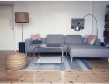 Wohnzimmer mit einer grauen Eckcouch, pastellfarbenen Kissen und einem Teppich auf einem Holzboden. Vor der Couch steht ein kleiner Metall- und Glastisch, daneben ein runder, geflochtener Pouf; im Hintergrund eine Stehlampe und Bücherregale.