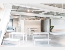 Heller, moderner Innenraum mit minimalistischer Einrichtung, darunter weiße Tische und Bänke, ein hochgelegener Schlafbereich mit Leiter und ein kleines Bar-Tisch-Setup mit schwarzen Hockern. Ein Topfpflanze steht in der Ecke, während der Raum durch helle Vorhänge und eine Beton-Decke ergänzt wird.