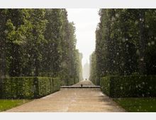 Ein gerader Weg in einem formalen Park, gesäumt von hohen, gepflegten Hecken und Bäumen, während Regen oder feiner Sprühregen fällt; im Hintergrund ist die Silhouette einer Kuppel oder eines historischen Gebäudes sichtbar.