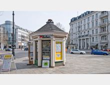 Ein historischer Verkaufsstand mit einem achteckigen Grundriss und Dachziegeln, an dem Zigaretten und Tickets verkauft werden, steht auf einem Bürgersteig einer städtischen Straße. Im Hintergrund sind ein modernes Glasgebäude und ein großes, verziertes Altbaugebäude mit Balkonen zu sehen.