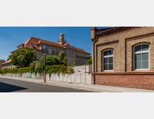 Straßenszene mit einem historischen Backsteingebäude rechts im Vordergrund und einem größeren Gebäude mit roten Ziegeldach und Uhrturm im Hintergrund. Eine bepflanzte Mauer mit Bäumen trennt die Gebäude von der Straße.