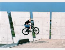 Ein Mann führt einen Sprung mit einem Mountainbike auf einer Betonrampe vor, während im Hintergrund eine Wand aus großen Betonplatten und ein klarer blauer Himmel zu sehen sind.