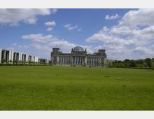 Das Bild zeigt den Reichstagsgebäude in Berlin, eine historische Regierungsgebäude mit Glaskuppel und klassizistischer Fassade. Im Vordergrund befindet sich eine große, offene Rasenfläche, umgeben von weiteren modernen Gebäuden und Bäumen unter einem leicht bewölkten Himmel.