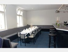 Ein kleiner Veranstaltungsraum mit einer langen, weiß gedeckten Tafel, Stühlen und einer gepolsterten Sitzbank entlang der Wand. Auf der rechten Seite befindet sich eine Bar mit hohen Barhockern, und mehrere Fenster lassen Tageslicht herein.