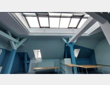 Ein moderner Dachraum mit blauen Wänden und sichtbaren weißen Stützbalken. Das Oberlicht mit großen Fenstern lässt Tageslicht herein, während einzelne Schreibtische und ein Regal den Raum möblieren.