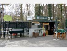 Außenansicht eines kleinen Imbissstands namens „Seehaase“ in einem bewaldeten Gebiet. Der Stand hat eine grüne Markise, Schilder für Getränke und Speisen sowie Tische und Stühle mit grünen Sonnenschirmen im Außenbereich.
