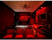 Ein kleiner, dunkel beleuchteter Veranstaltungsraum mit roten Wänden und einem roten Sofa im Vintage-Stil. Im Hintergrund ist eine Bühne mit einem Wandbild und schwacher Beleuchtung zu sehen, umgeben von Vorhängen und dekorativen Möbeln.