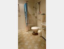 Badezimmer mit Fliesenboden in Beige, einer Duschkabine mit Glaswand und Duschvorhang, einer Toilette, einem kleinen Mülleimer sowie einem Waschbecken mit Unterschrank. Die Wände sind hell gefliest.