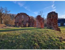 Ruine eines Backsteingebäudes mit gotischen Bögen und verbliebenen Wänden, umgeben von einer grünen Wiese und Bäumen im Hintergrund; rechts außen ist ein Fachwerkgebäude erkennbar. Der Himmel ist klar und blau.
