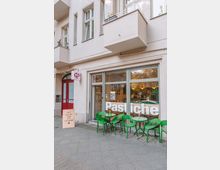 Street-level view of a cafe storefront in a light stucco apartment building, with large front windows labeled “Pastiche” and a small sidewalk seating area of green chairs and round tables. A red entry door and a small hanging sign are beside the windows, with balconies above.