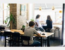 Ein modernes Café mit großen Fenstern, Ziegelwänden und Holztischen. Im Vordergrund sitzt eine Person an einem Tisch mit Laptop, während im Hintergrund zwei Personen an einer Bartheke nahe der Fensterfront sitzen.