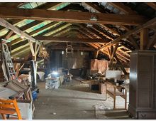 Dachbodenraum mit sichtbaren Holzbalken und einem schrägen Dach, gefüllt mit gelagerten Möbeln, Kartons und Gegenständen. Die Umgebung ist schwach beleuchtet, und Tageslicht fällt durch das Dach und kleine Fenster.