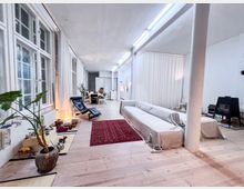 Ein geräumiger Wohnraum mit hellen Holzböden, großen Sprossenfenstern und weißen Wänden. Der Raum ist mit einer langen beigefarbenen Couch, einem roten Teppich, Pflanzen und modernen Möbeln eingerichtet; weiße Vorhänge und freistehende Säulen teilen den offenen Raum.