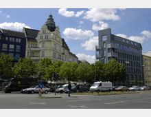 Straßenszene mit mehrstöckigen Gebäuden im Hintergrund, darunter ein hellgelbes historisches Gebäude mit Turm und ein modernes dunkelgraues Gebäude mit Außentreppen. Vordergrund mit parkenden Autos, einem Lieferwagen und einem Fußgänger an einer Ampel, umgeben von Bäumen.