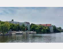 Flussufer mit Gebäuden im Hintergrund, darunter mehrstöckige Wohnhäuser mit roten und grauen Dächern. Am Ufer befinden sich viele grüne Bäume, einige davon sind Weiden, sowie ein kleiner Bereich mit weißen Sonnenschirmen.