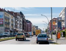 Straßenszene in einer deutschen Stadt mit mehrstöckigen Wohn- und Geschäftsgebäuden entlang einer befahrenen Straße. Im Vordergrund parken und fahren Autos, rechts befindet sich ein Gehweg mit einer Tankstellenpreistafel und einer orangenen Postbox.