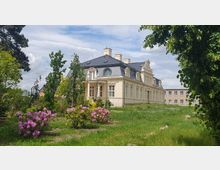 Historisches Gebäude mit cremefarbener Fassade und Mansardendach, umgeben von einer grünen Gartenanlage mit blühenden Sträuchern und Bäumen; im Hintergrund befindet sich ein einfaches, modernes Gebäude.