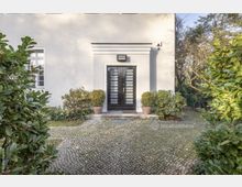 Helle Fassade eines Wohnhauses mit einer mittig angeordneten, schwarzen Eingangstür. Der gepflasterte Bereich vor der Tür ist von Pflanzen und Sträuchern in Töpfen sowie im Garten gesäumt, im Hintergrund sind Bäume sichtbar.