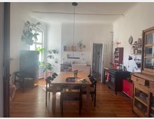 Helle Wohnküche mit Holzboden, großem Esstisch und schwarzen Stühlen. An den weißen Wänden befinden sich offene Regale und Pflanzen, während Möbel aus Holz sowie ein schwarzer Herd das Kücheninterieur bestimmen.