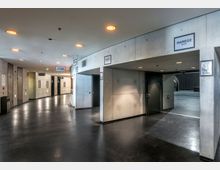 Moderner Innenbereich mit glatten Betonwänden, dunklem Boden und eingebauten Deckenspots. Mehrere Türen, darunter Eingänge mit Beschilderung wie "Manege Mitte" und Wegweiser zu WC, Lift und anderen Räumen, prägen den Flur.