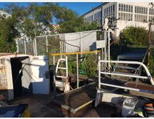 Eine kleine, rustikale Plattform mit Metallgeländern, Holzplanken und einer Zugangstür, umgeben von grünen Büschen und einem hohen Zaun. Im Hintergrund sind ein weißes Industriegebäude mit großen Fenstern und ein klarer blauer Himmel zu sehen.