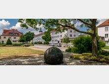 Eine kleine Stadtplatzszene mit einem gepflasterten Weg, einer großen Stein-Skulptur in Kugelform im Vordergrund und mehreren Wohngebäuden mit roten Ziegeldächern im Hintergrund. Links und rechts des Platzes befinden sich Grünflächen mit Bäumen und Sträuchern, im Hintergrund ist ein Kirchturm sichtbar.