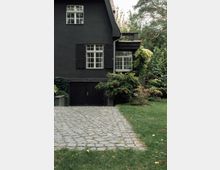Schwarzes Haus mit zwei Sprossenfenstern und Fensterläden, neben einer begrünten Terrasse. Im Vordergrund führt eine mit Pflastersteinen belegte Einfahrt durch einen Garten mit Bäumen und Sträuchern.