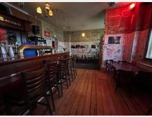 Innenraum einer schummrigen Kneipe mit langer Holztheke und Barhockern links, Holzdielenboden und Tischen mit Stühlen rechts. Die Wände sind dicht mit Graffiti und Kritzeleien bedeckt, beleuchtet von warmen Deckenlampen und rotem Licht im vorderen Bereich.