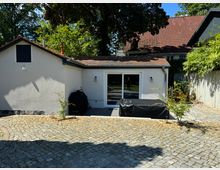 Einfamilienhaus mit weißer Fassade und roten Dachziegeln, davor ein gepflasterter Hof. Im Vordergrund stehen ein überdachter Grill und kleine Pflanzen, die an der Seite eines begrünten Gartens positioniert sind.