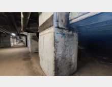 Langer, unterirdischer Betonflur mit rohen Wänden und sichtbaren Rohren an der Decke. Der Bereich ist schwach beleuchtet und zeigt Spuren von Abnutzung an den Oberflächen.