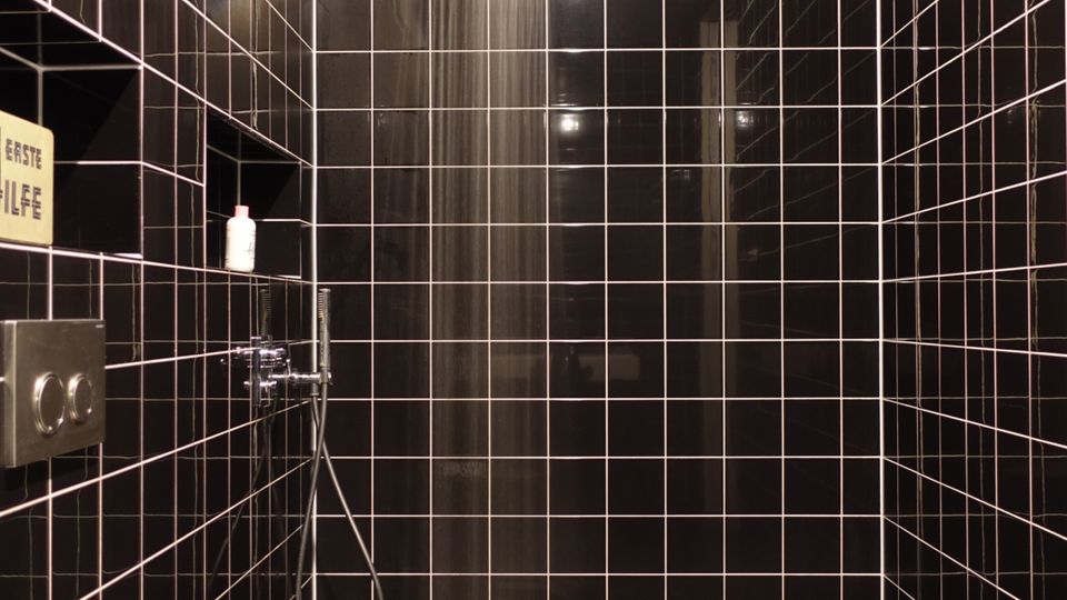 Ein modernes Badezimmer mit schwarzen Fliesen an Wänden und Boden. Zu sehen sind eine offene Regendusche mit herabhängender Brause und ein wandmontiertes WC.