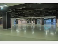 Großer, leerer Innenraum mit glänzendem Boden, vielen dunklen Deckenstrukturen und rechteckigen Säulen. Im Hintergrund befinden sich Glaswände und Fenster, die teilweise Tageslicht hereinlassen.