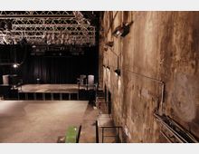 Industriehalle mit Bühne: Der Raum hat eine rohe Betonwand mit sichtbaren Rohren und Kabeln, eine einfache Bühne mit schwarzen Vorhängen im Hintergrund sowie Traversensysteme mit Beleuchtung und Lautsprechern an der Decke. Der Boden ist aus Beton, und der Raum wirkt funktional und minimalistisch.