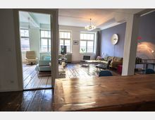 Wohnzimmer in einer Altbauwohnung mit Holzdielenboden und großen Fensterfronten, die viel Tageslicht hereinlassen. Der Raum ist mit einer Sitzecke, einem Holztisch, bunten Möbeln und einem minimalistischen Lampen-Design eingerichtet.