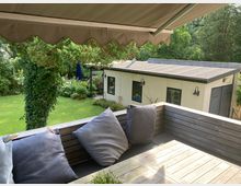 Terrasse mit Holzsitzbank und Kissen im Vordergrund, Sonnenschutz oben. Dahinter ein gepflegter Garten mit Rasen, Bäumen und einem weißen Gartenhaus mit flachem Dach und dunklen Fenstern.