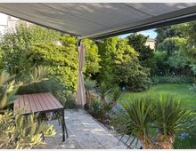 Überdachte Gartenterrasse mit Steinplatten, Esstisch und Stühlen unter einer Markise mit seitlichem Vorhang. Daneben ein gepflegter Rasen und dichter Gartenbewuchs mit Sträuchern, Nadelbäumen und palmenähnlichen Pflanzen entlang der Beetkante.